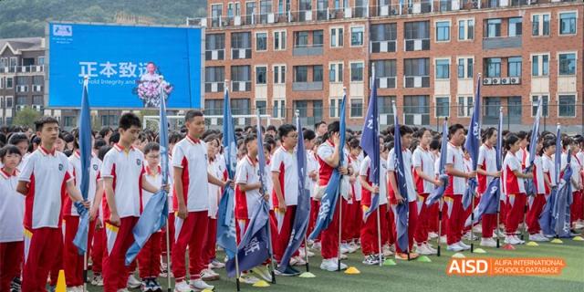 Guiyang Prime School students standing in formation outdoors, holding flags or banners during a school event or sports day, with school buildings in the background.