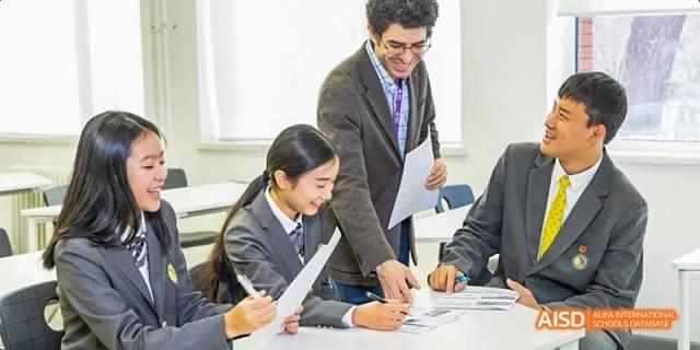 An educator smiles while assisting a female student with her coursework at a group desk in Beijing Haidian Kaiwen Academy.