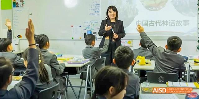 A female teacher stands at the front of a modern classroom gesturing while speaking to seated students at Beijing Haidian Kaiwen Academy.
