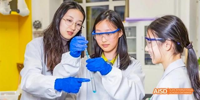 Two female students wearing lab coats and safety goggles conduct a chemistry experiment with a pipette at Beijing Haidian Kaiwen Academy.