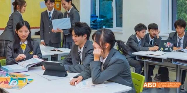 Students in formal grey uniforms collaborate using a tablet and notebook during a group study session at Beijing Haidian Kaiwen Academy.