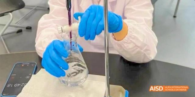 A close-up of a student's gloved hands carefully pouring a blue liquid into a glass flask during lab at Beijing Haidian Kaiwen Academy.