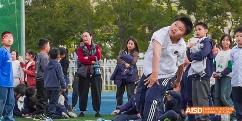 A student in a white shirt lunges forward during an outdoor physical activity on a blue court with onlookers in the background at Beijing Haidian Kaiwen Academy.