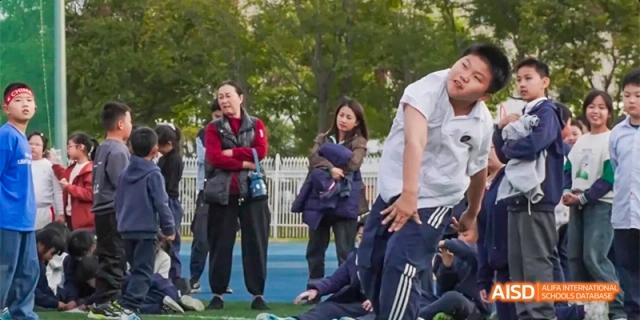 A student in a white shirt lunges forward during an outdoor physical activity on a blue court with onlookers in the background at Beijing Haidian Kaiwen Academy.
