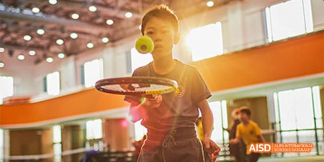 A young student focuses on a yellow tennis ball hovering near his racket during an indoor practice session bathed in sunlight at Beijing Haidian Kaiwen Academy.