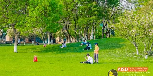 Students and families relax and sit on a lush green grassy hill under the shade of tall trees during a sunny outdoor event at Beijing Haidian Kaiwen Academy.