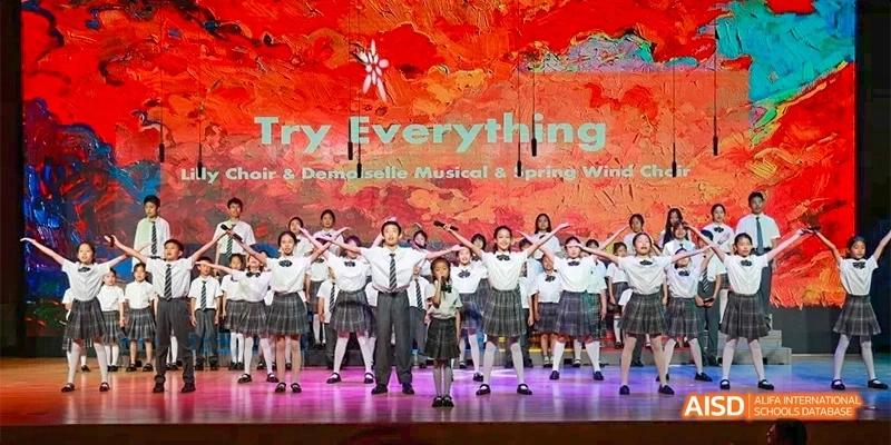 A large student choir in school uniforms performs "Try Everything" on a stage with a vibrant red and orange backdrop at Beijing Haidian Kaiwen Academy.