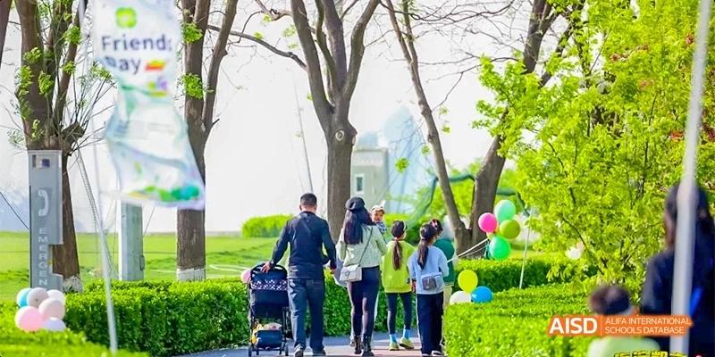 A family walks together along a paved campus path lined with green hedges and colorful balloons during a bright spring day at Beijing Haidian Kaiwen Academy.