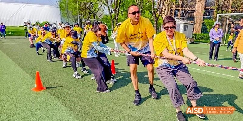 A team of staff and students in yellow shirts work together during a high-energy tug-of-war competition on an outdoor field at Beijing Haidian Kaiwen Academy.