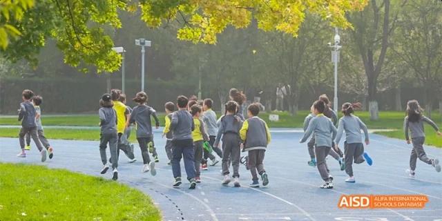 A large group of students in grey and yellow uniforms jogging together on a blue outdoor athletic track during a physical education class at Beijing Haidian Kaiwen Academy.