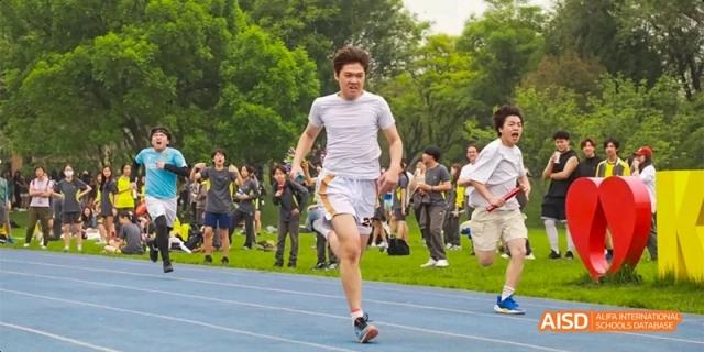 Students competing in an intense relay race on a blue track, with a runner in white sprinting forward while others cheer in the background at Beijing Haidian Kaiwen Academy.