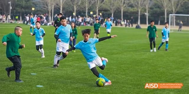 A young student in a blue jersey skillfully dribbling a soccer ball across a lush green grass field during a competitive match at Beijing Haidian Kaiwen Academy.