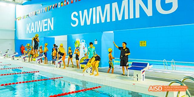 Students in swimsuits and caps lined up along the edge of a bright blue indoor swimming pool while an instructor gives directions at Beijing Haidian Kaiwen Academy.