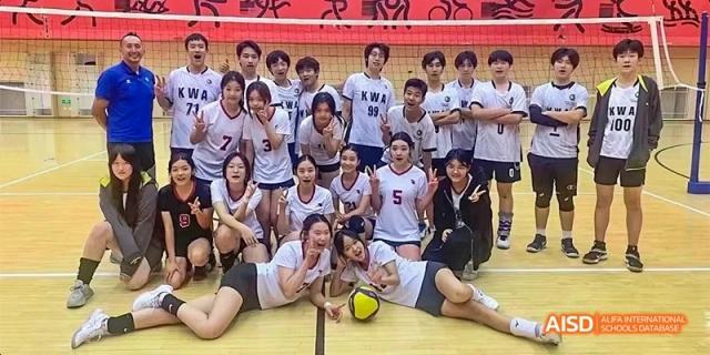 A large, smiling co-ed volleyball team posing together on an indoor court, wearing white and maroon jerseys after a successful game at Beijing Haidian Kaiwen Academy.