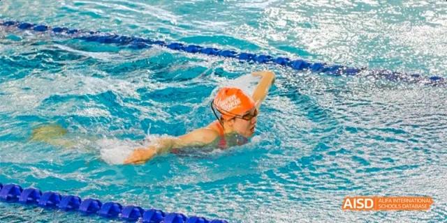 A student athlete wearing an orange swim cap performing a freestyle stroke during a swim meet in a professional indoor competition pool at Beijing Haidian Kaiwen Academy.