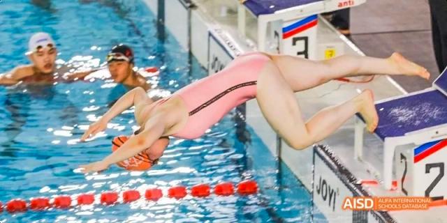 A female swimmer in a pink swimsuit captured mid-air while diving off a starting block into the water at the start of a race at Beijing Haidian Kaiwen Academy.