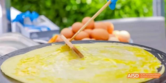 A chef uses a wooden spreader to thin out batter on a large flat griddle to create fresh crepes for students at Beijing Haidian Kaiwen Academy.