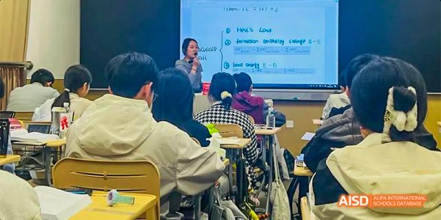 A teacher stands at the front of a classroom by a whiteboard filled with chemistry notes, instructing a focused group of students at Hefei No.8 High School.