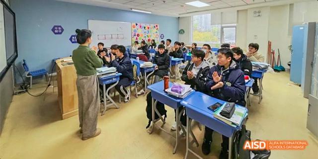 A group of students sit at blue desks in a classroom, clapping and engaging with a peer standing at the front during a lesson at Hefei No.8 High School.
