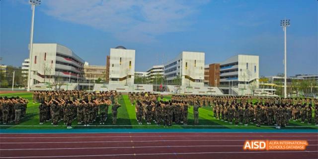 A large group of students in military uniforms standing on a green field in front of the school building at Hefei No.8 High School.