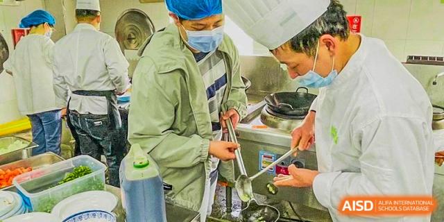 A student in a hairnet and mask learning to use a large ladle in a kitchen at Hefei No.8 High School.