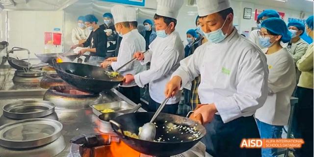 A line of students in chef hats and masks practicing stir-frying with large woks at Hefei No.8 High School.