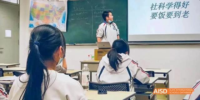 A student from Kunming No.1 High School International Education is seated at a desk while a teacher stands nearby, offering assistance.