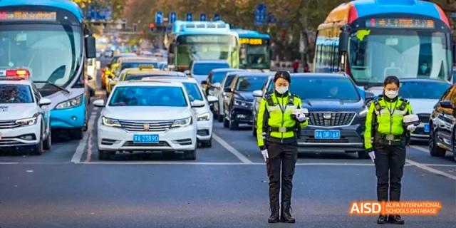 A police officer from Kunming No.1 High School International Education directs traffic in a busy street, with cars and buses in the background.