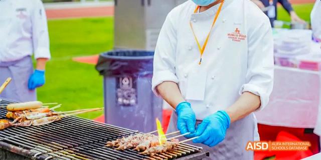 A chef from Lady Eleanor Holles International School Foshan(LEH Foshan) wearing blue gloves cooks meat skewers on a grill during an outdoor event.
