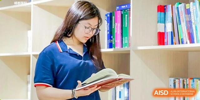 A female student with glasses reading a book while standing next to a bookshelf at Orange County American High School Beijing Campus.