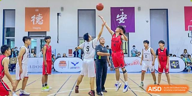 A student attempting a jump shot in a basketball game while being defended, with a referee nearby, at Orange County American High School Beijing Campus.