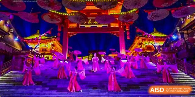 A cultural performance with people in traditional dress, holding umbrellas on a brightly lit stage at Orange County American High School Beijing Campus.