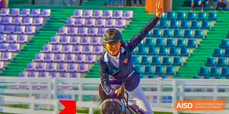 A student participating in an equestrian event, smiling and raising their hand while riding a horse at Orange County American High School Beijing Campus.