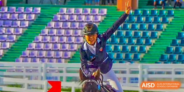 A student participating in an equestrian event, smiling and raising their hand while riding a horse at Orange County American High School Beijing Campus.