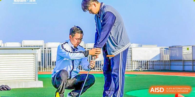 A coach or instructor kneeling to adjust a student's gear or measurement on an outdoor track at Orange County American High School Beijing Campus.