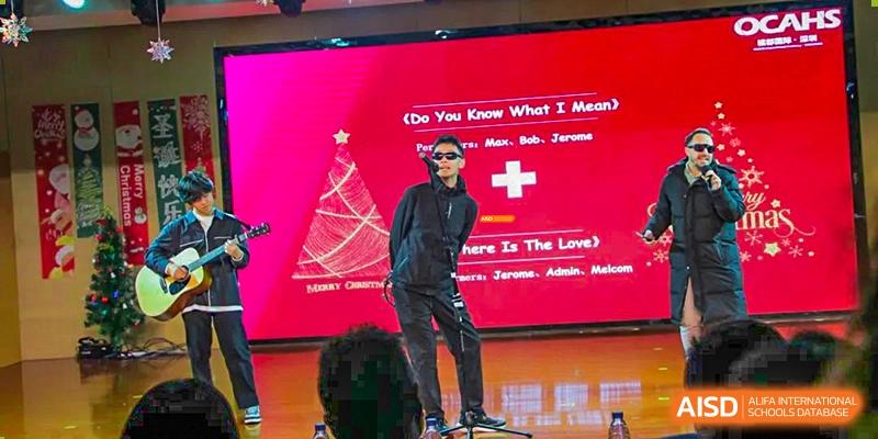 A person in sunglasses and all-black attire performing on a stage with a red Christmas tree graphic in the background at Orange County American High School Beijing Campus.