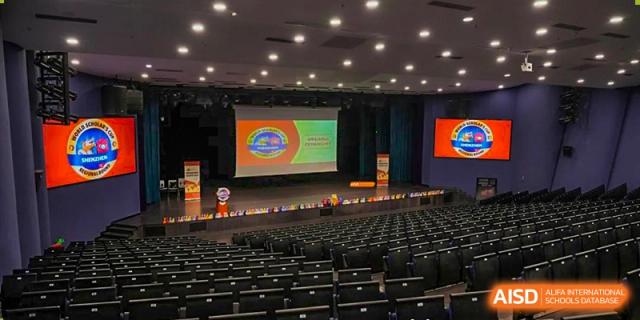 A wide-angle view of a large, empty auditorium or theatre with rows of seating and a brightly lit stage at Orange County American High School Beijing Campus.
