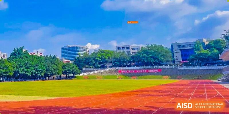 A bright orange frisbee flying over a green and red outdoor sports field with school buildings in the background at Orange County American High School Beijing Campus.
