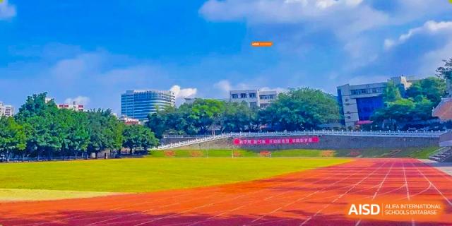 A bright orange frisbee flying over a green and red outdoor sports field with school buildings in the background at Orange County American High School Beijing Campus.