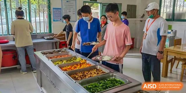 Two male students using tongs to select food from a buffet line with hot dishes and vegetables in the school cafeteria at Orange County American High School Beijing Campus.