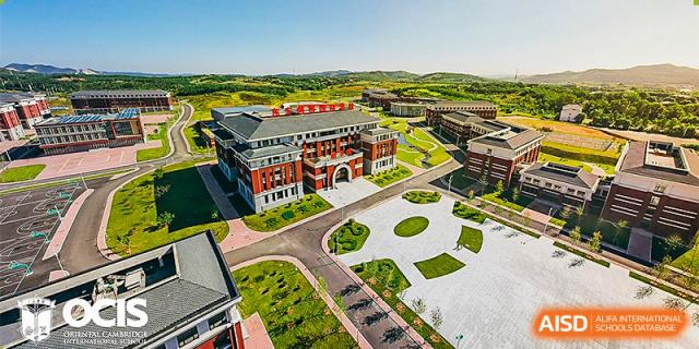An aerial view of the main courtyard and academic buildings of Oriental Cambridge International School, showing the red brick architecture and surrounding greenery.