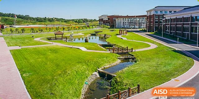 An aerial view of the green campus grounds of Oriental Cambridge International School, featuring landscaped lawns, streams, and bridges.