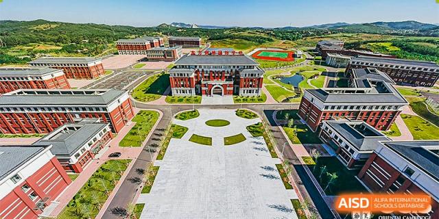 A wide aerial view showing the extensive main plaza and red brick academic buildings of Oriental Cambridge International School, surrounded by rolling green hills.