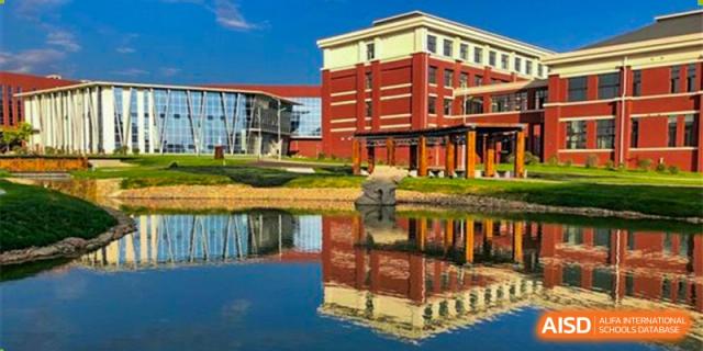 An exterior view of the large red brick building and glass facade of Oriental Cambridge International School, with a reflection in the adjacent pond.