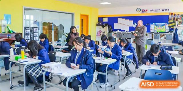 Students at Oriental Cambridge International School in school uniform seated at desks in a classroom, focusing on their work and a book.
