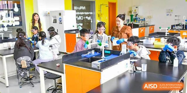 Students and a teacher at Oriental Cambridge International School wearing gloves and lab coats while conducting an experiment at a sink in a brightly colored science lab.