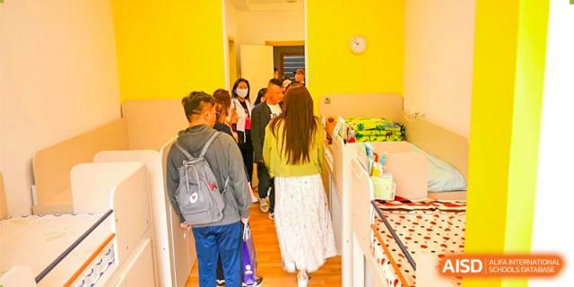 A group of parents or visitors interacting with staff and students in a brightly lit, yellow-walled classroom at Oriental Cambridge International School.