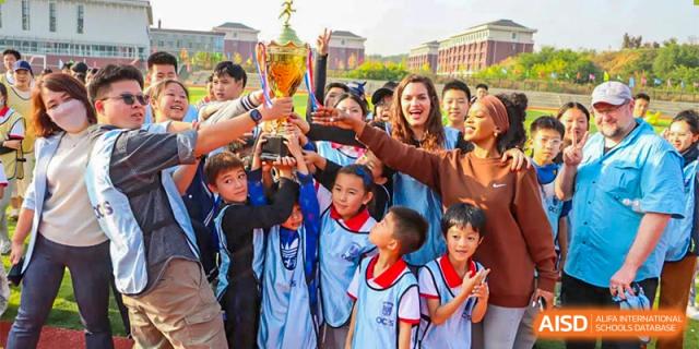 A group of diverse students and adults at Oriental Cambridge International School smiling and posing for a group photo, with several hands holding up cameras or phones.