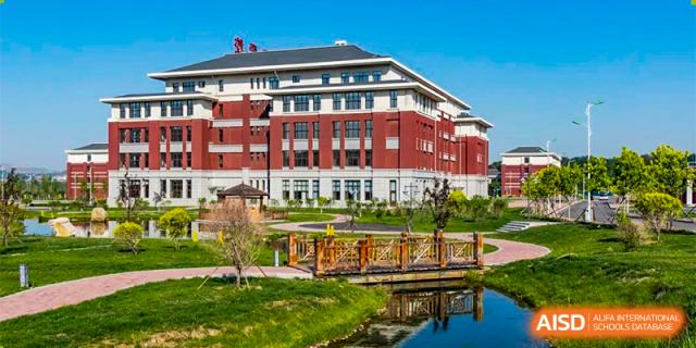 An exterior view of the red brick building on the campus of Oriental Cambridge International School, with a small wooden bridge over a stream or pond in the foreground.