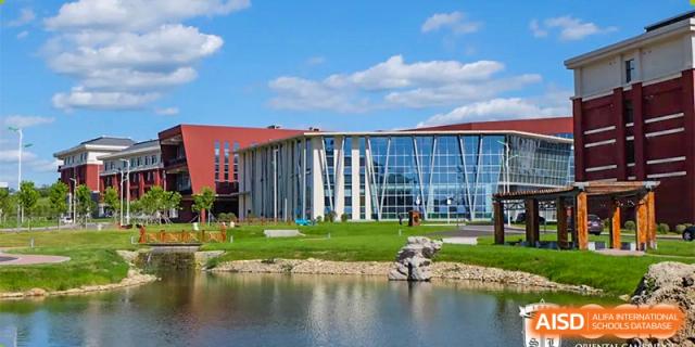 An exterior view of the modern campus of Oriental Cambridge International School, showing a large glass-fronted building and a man-made pond in the foreground.